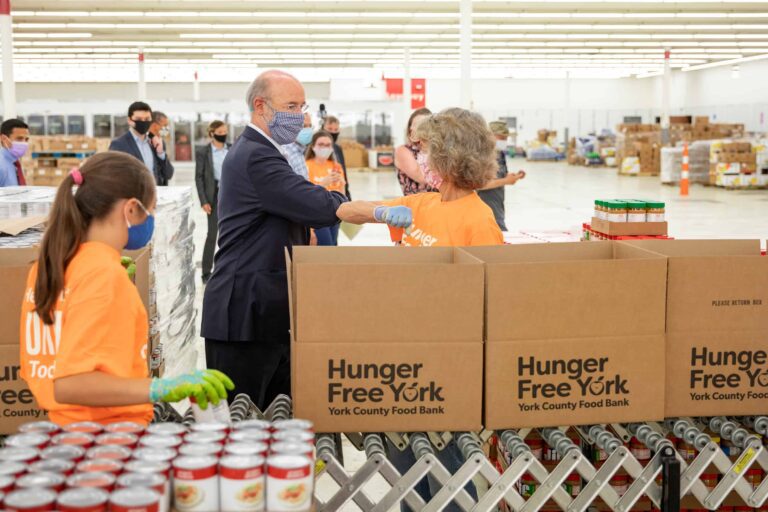 An older man bumps elbows with a woman standing by boxes of food.