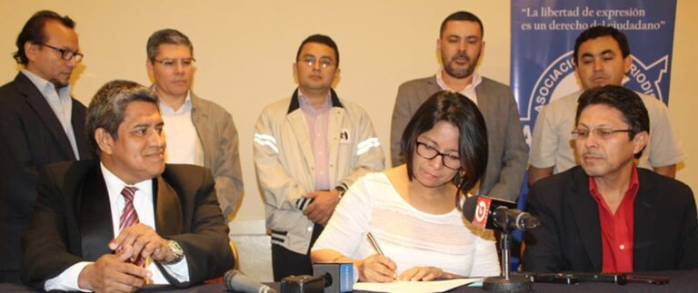 A group of men sit and stand around a woman who is writing on a paper.