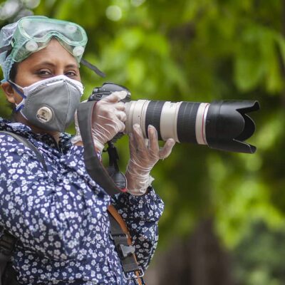A woman wearing goggles, a face mask, and gloves holds a large camera
