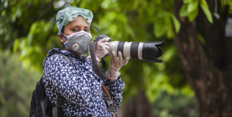 A woman wearing goggles, a face mask, and gloves holds a large camera