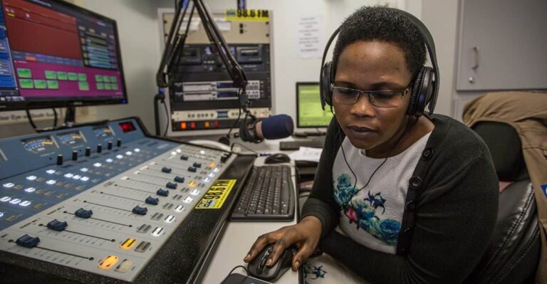 A woman wearing headphones sits in front of a mixing board in a radio studio