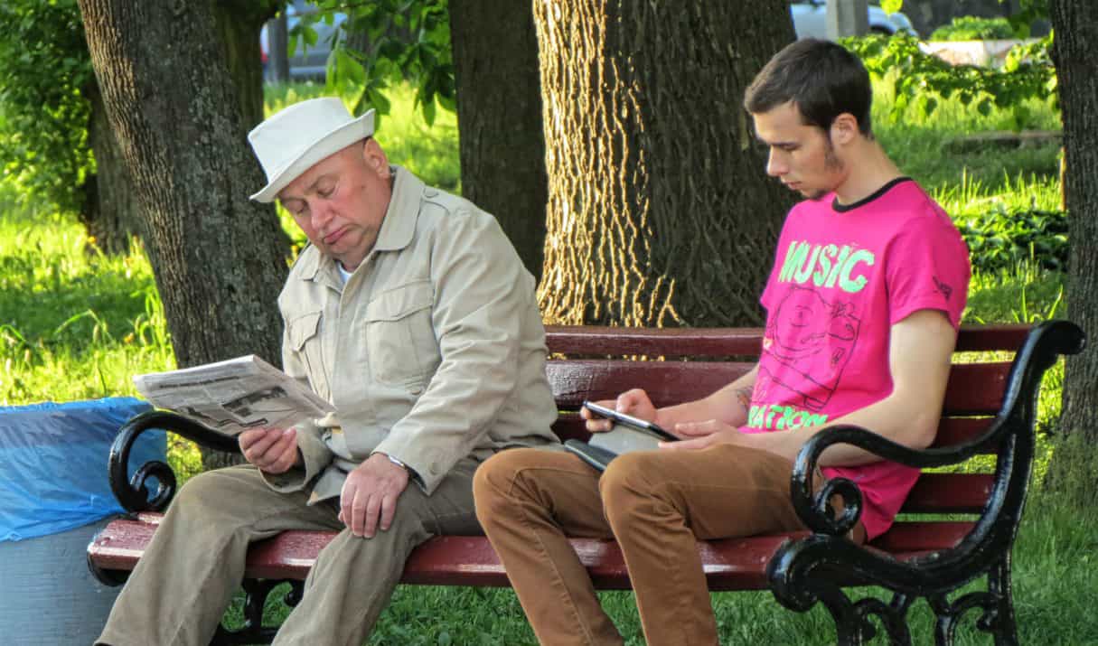 Two men sit on a bench; the older man reads a newspaper; the young man reads an tablet.