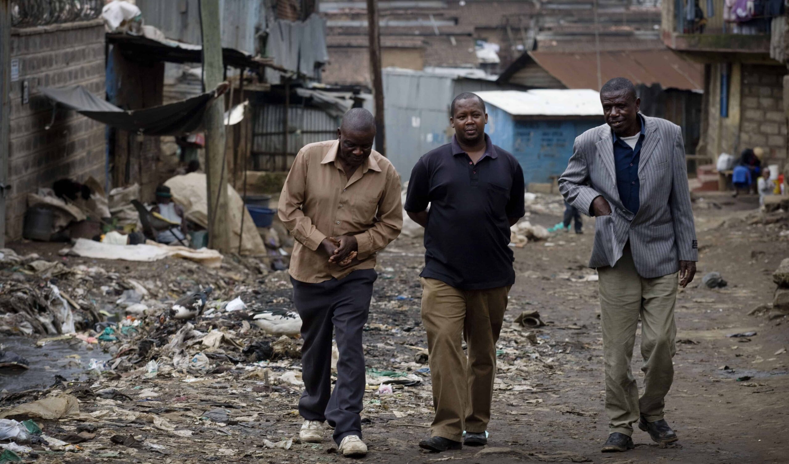 Three men walk down a garbage strewn dirt road.