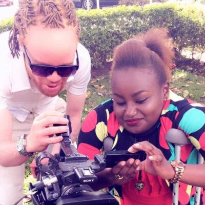 A light skinned man with braided hair and a dark skinned woman on crutches look at a video camera