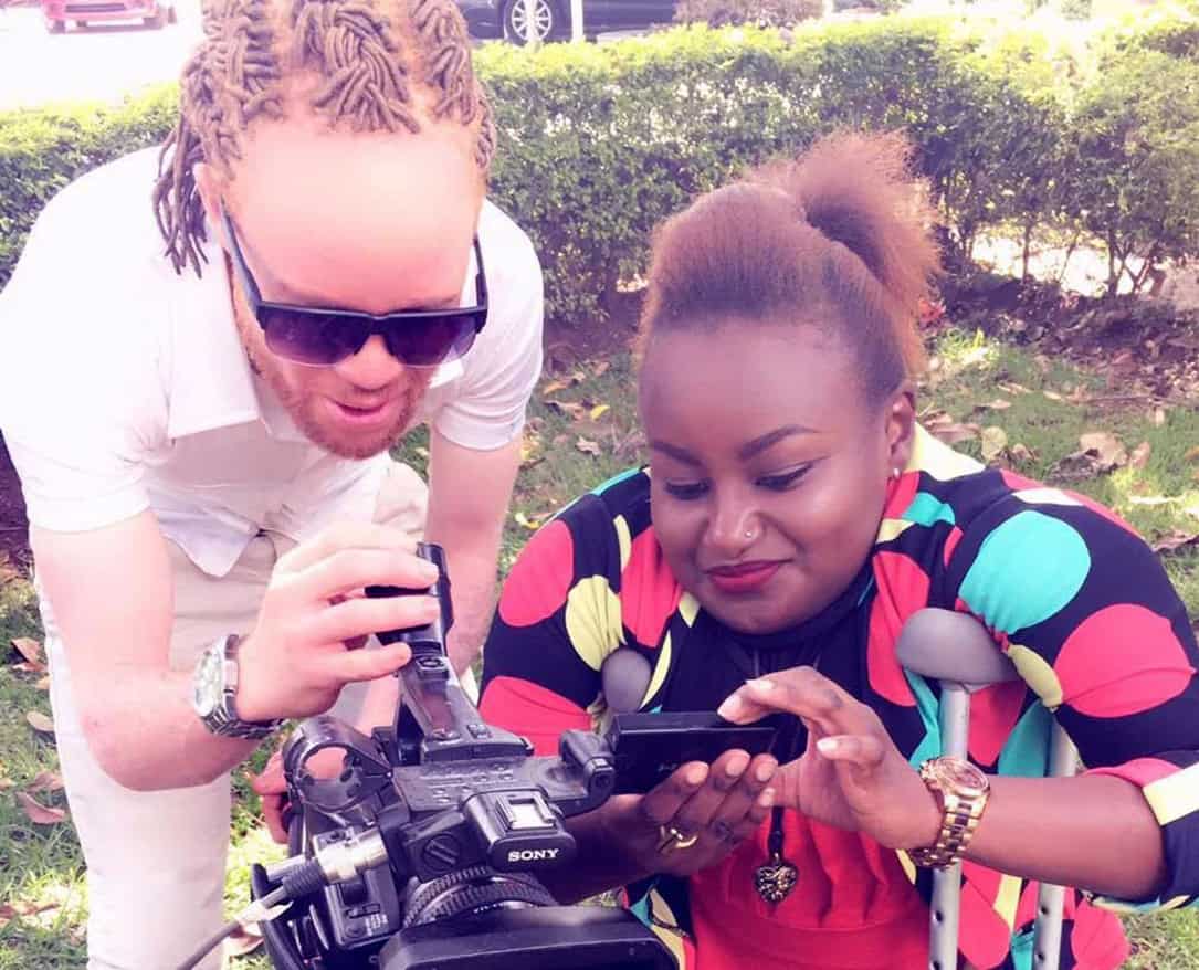 A light skinned man with braided hair and a dark skinned woman on crutches look at a video camera