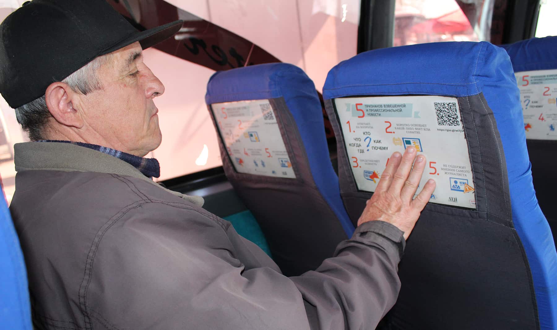A man sitting on a bus reads the poster mounted on the seat in front of him.