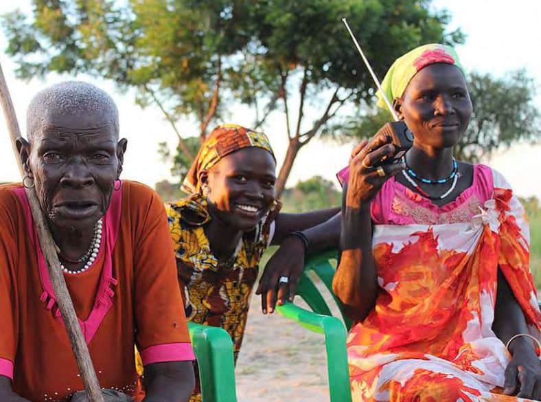 An older woman and two younger women sit outside listening to a portable radio.