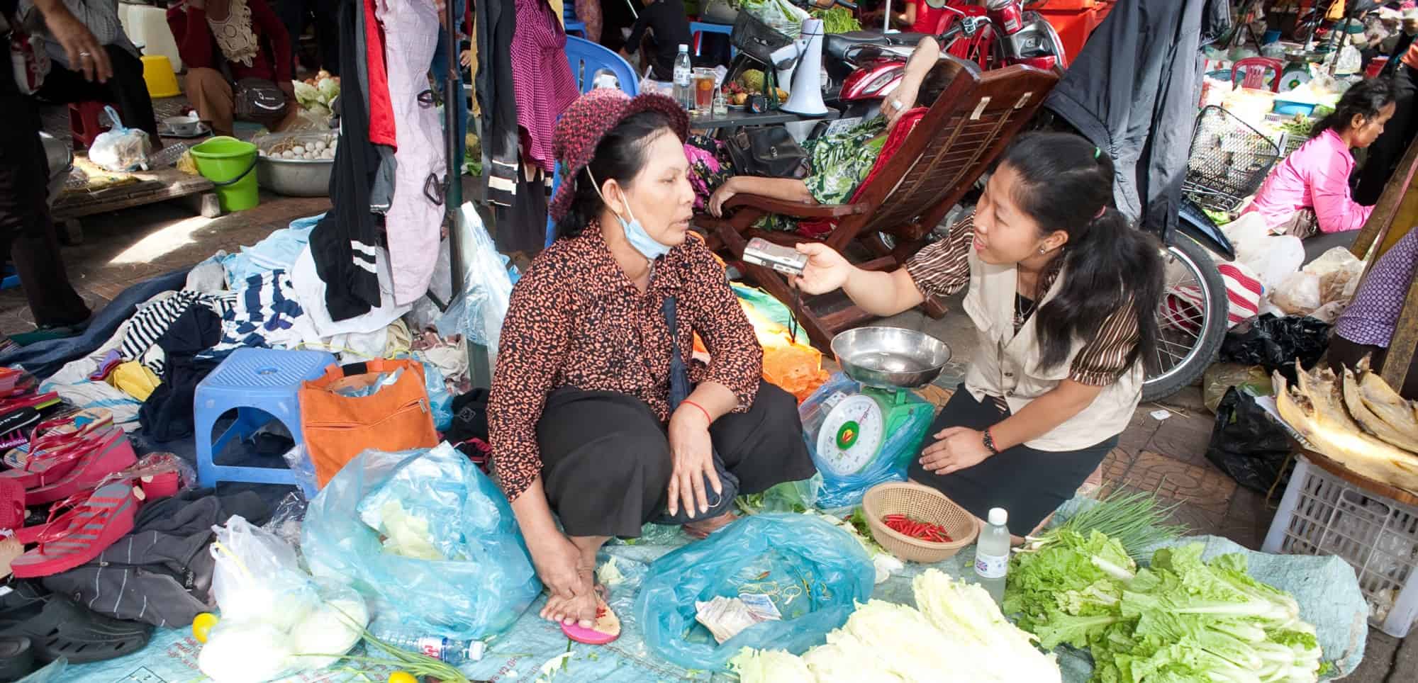 A journalist interviews a woman selling vegetables in a market.