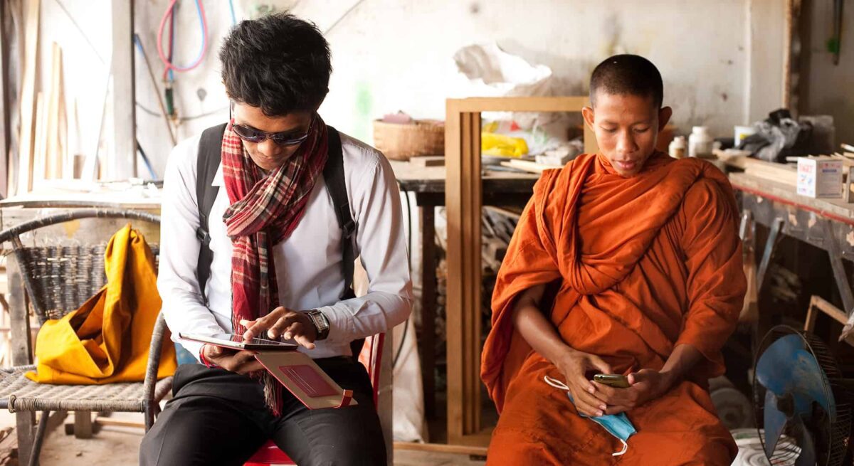 A man in a white shirt, dark pants, sunglasses, and wearing a backpack sits working on a tablet. Another man sits next to him, wearing an orange robe and looking at a smart phone.