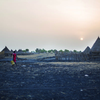 A woman walks through a dirt field by thatched roof structures; she is carrying a metal container.
