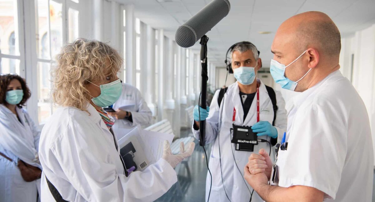 A woman wearing a face mask talks to a man wearing a face mask, while another man records them on a mic