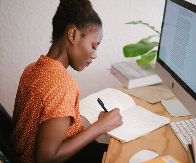 A woman writes in a notebook while sitting in front of a monitor.