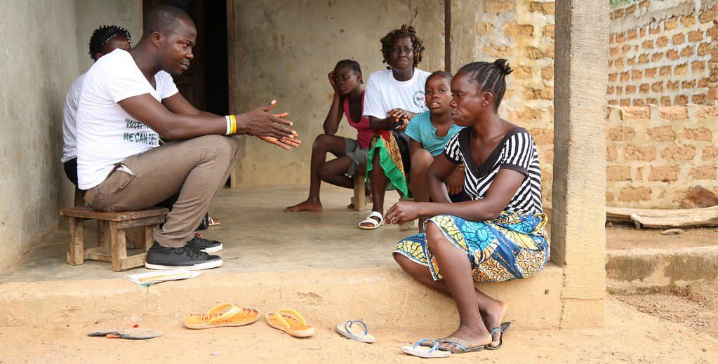 A man sits with a group of women and children on a concrete patio outside a brick building