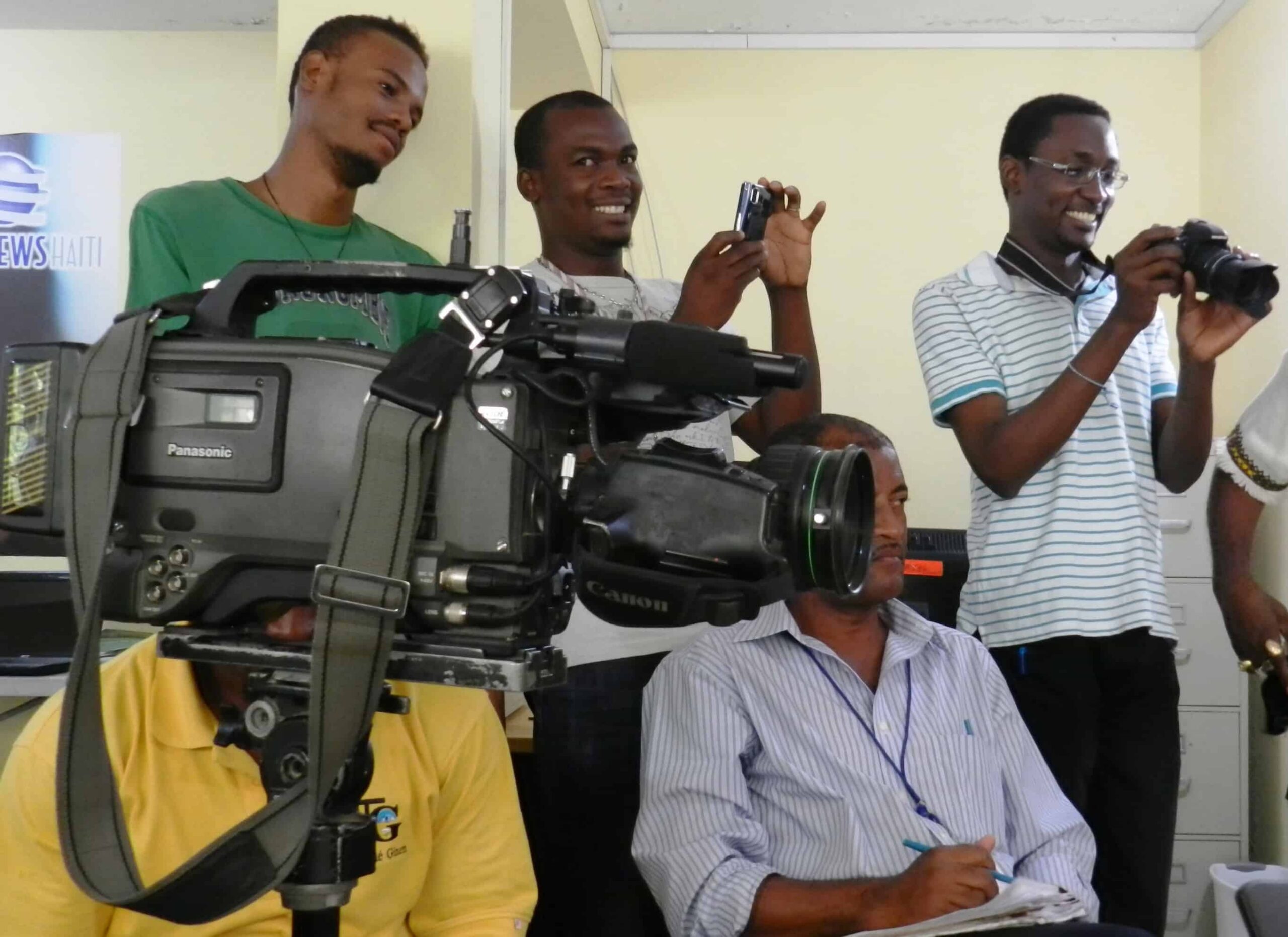 Five men stand in a room; some using smart phones; one has a video camera.
