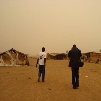 Two men walk on the dirt ground among some shelters constructed of sticks, reeds, and fabric.