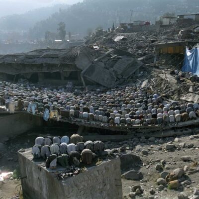 A few hundred people bow in prayer by a concrete building destroyed by an earthquake