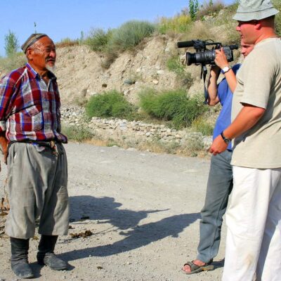 A man stands on a dirt road in a desert being filmed by two videographers.