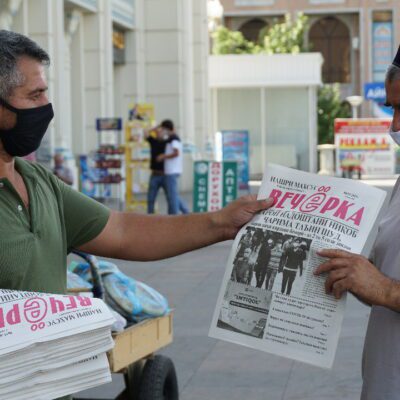 A man wearing a face covering hands a newspaper to another man