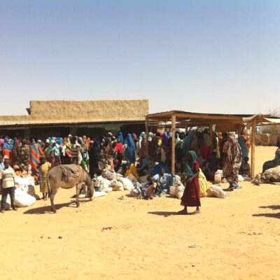 A large group of people stand under a shelter in the desert. Many have large sacks.