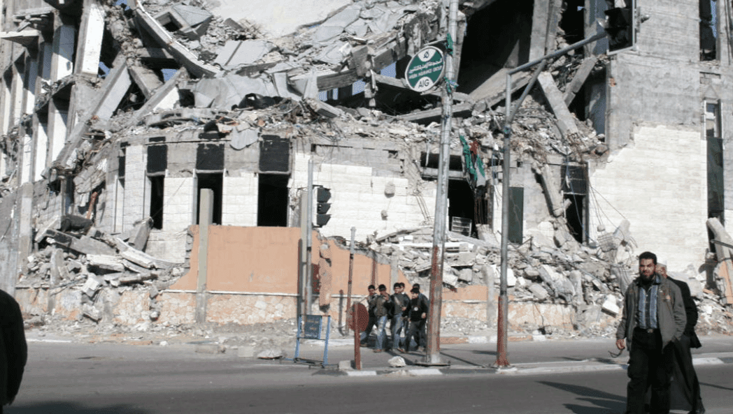 People walk in the street by a building damaged by bombing