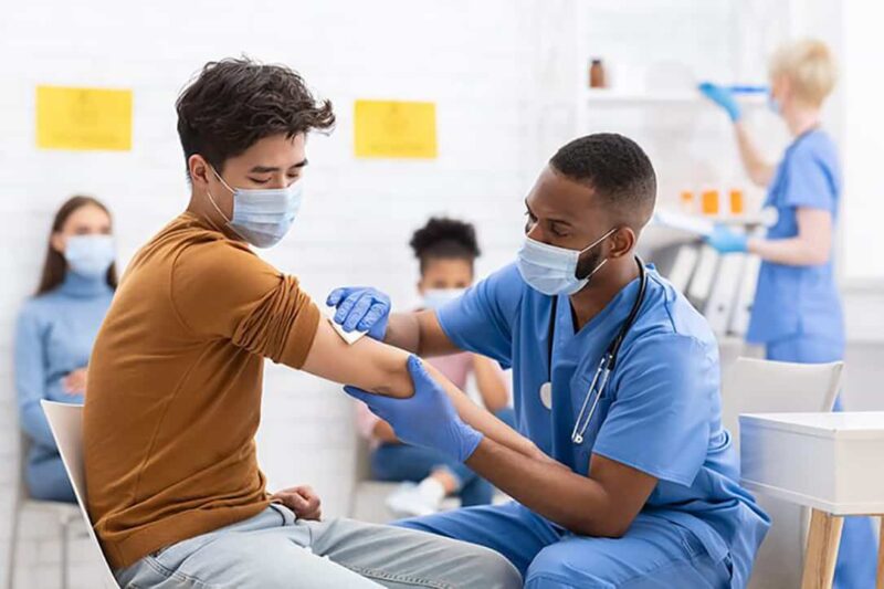 A man wearing blue scrubs and a face mask gives an injection to a man.