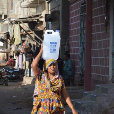 A woman walks down a street carrying a large plastic bottle on her head.