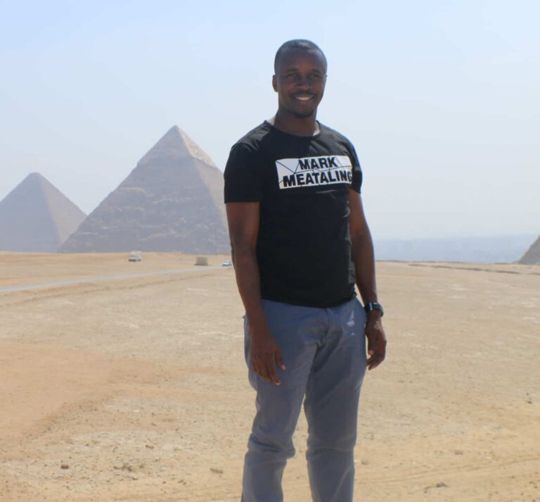 A man stands in the desert with pyramids in the background
