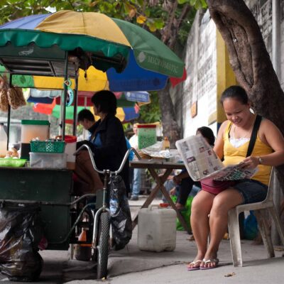A woman sits on a chair on the street reading a newspaper; she's next to a food cart.