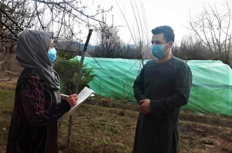 A woman holding a pen and a pad of paper talks to a man; both are wearing face masks.
