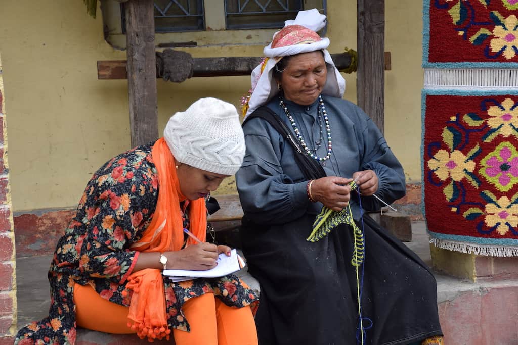 Two women sit together; one is knitting; the other is writing on a notepad.