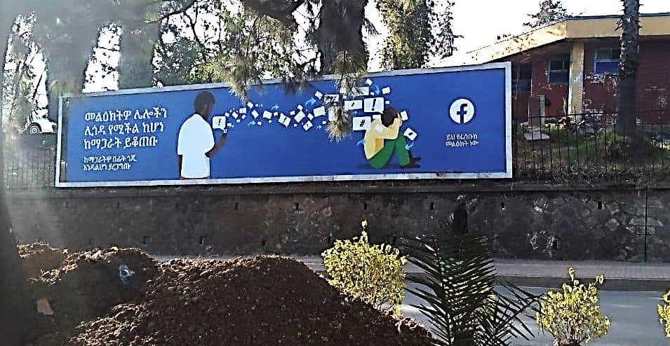 A billboard with Amharic script and a Facebook logo along side of a road.
