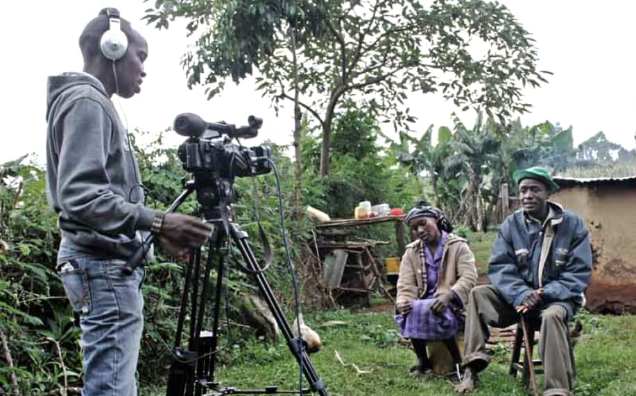 A man stands behind a video camera on a tripod taping a woman and a man who are both seated.
