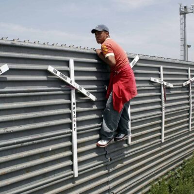 A man holds on to the side of a tall fence. White crosses are attached to the fence.