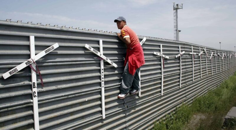 A man holds on to the side of a tall fence. White crosses are attached to the fence.