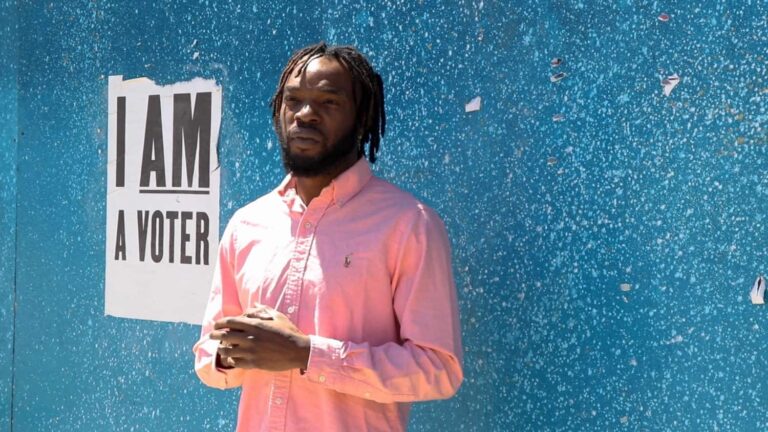 An African American man stands next to a poster that says "I am a Voter"