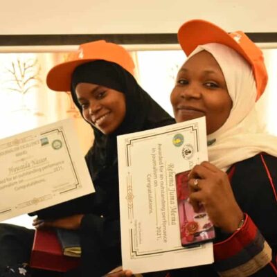 Two women, both wearing orange caps, hold certificates in their hands.