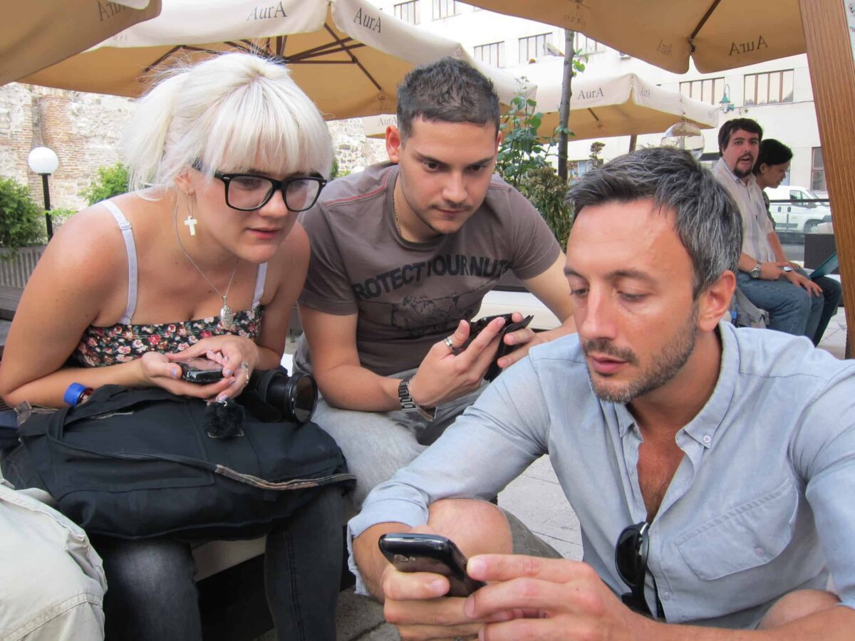 Two men and a woman wearing glasses sit together with their cell phones. The woman and young man look on as the other man shows something on his phone.