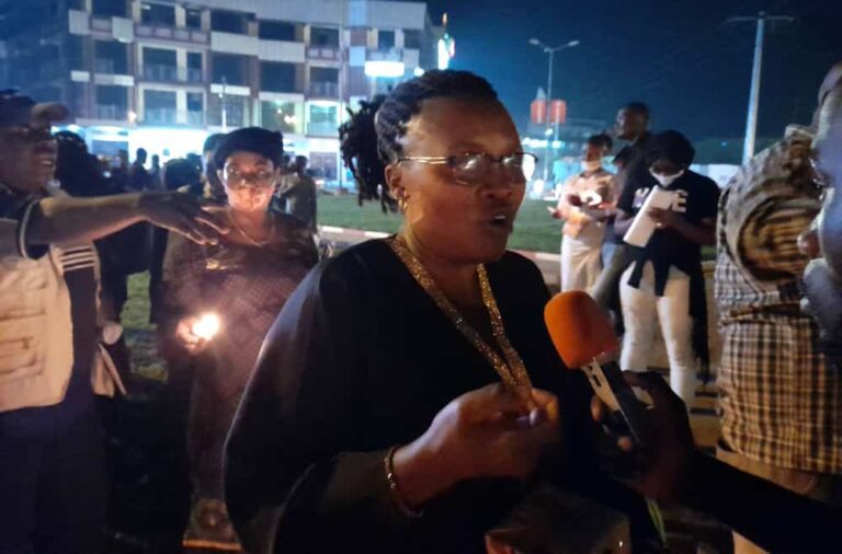 A group of people stand outside at night; some are holding candles; one woman speaks into a mic.