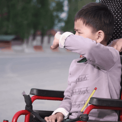 A boy sits in a wheelchair with someone standing behind him.