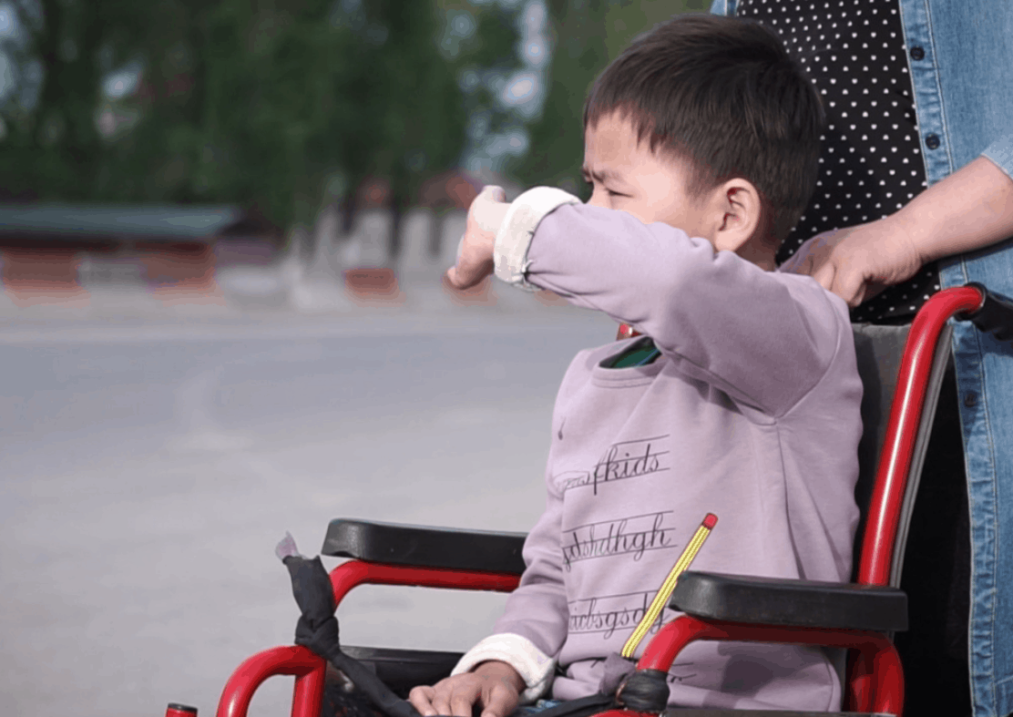A boy sits in a wheelchair with someone standing behind him.