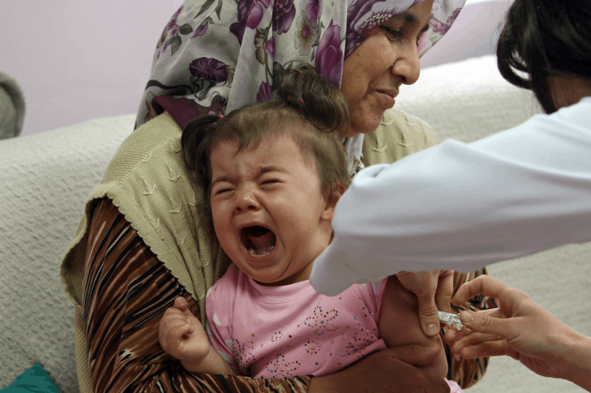 A woman holds a crying baby in her lap while another person administers a shot.