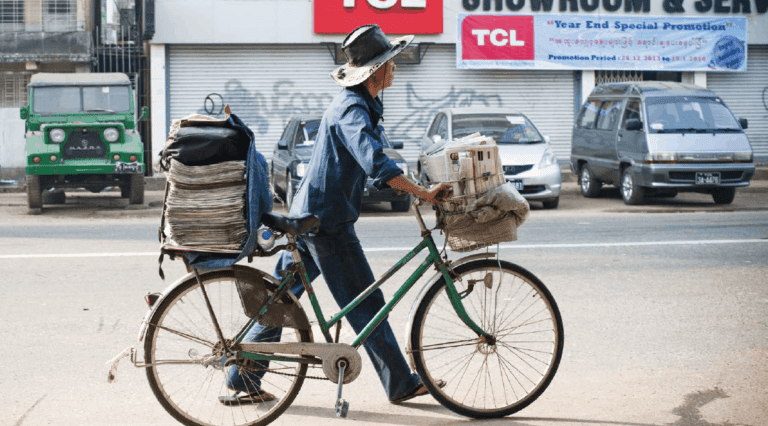 A man walks his bicycle, piled with newspapers, down a city street.