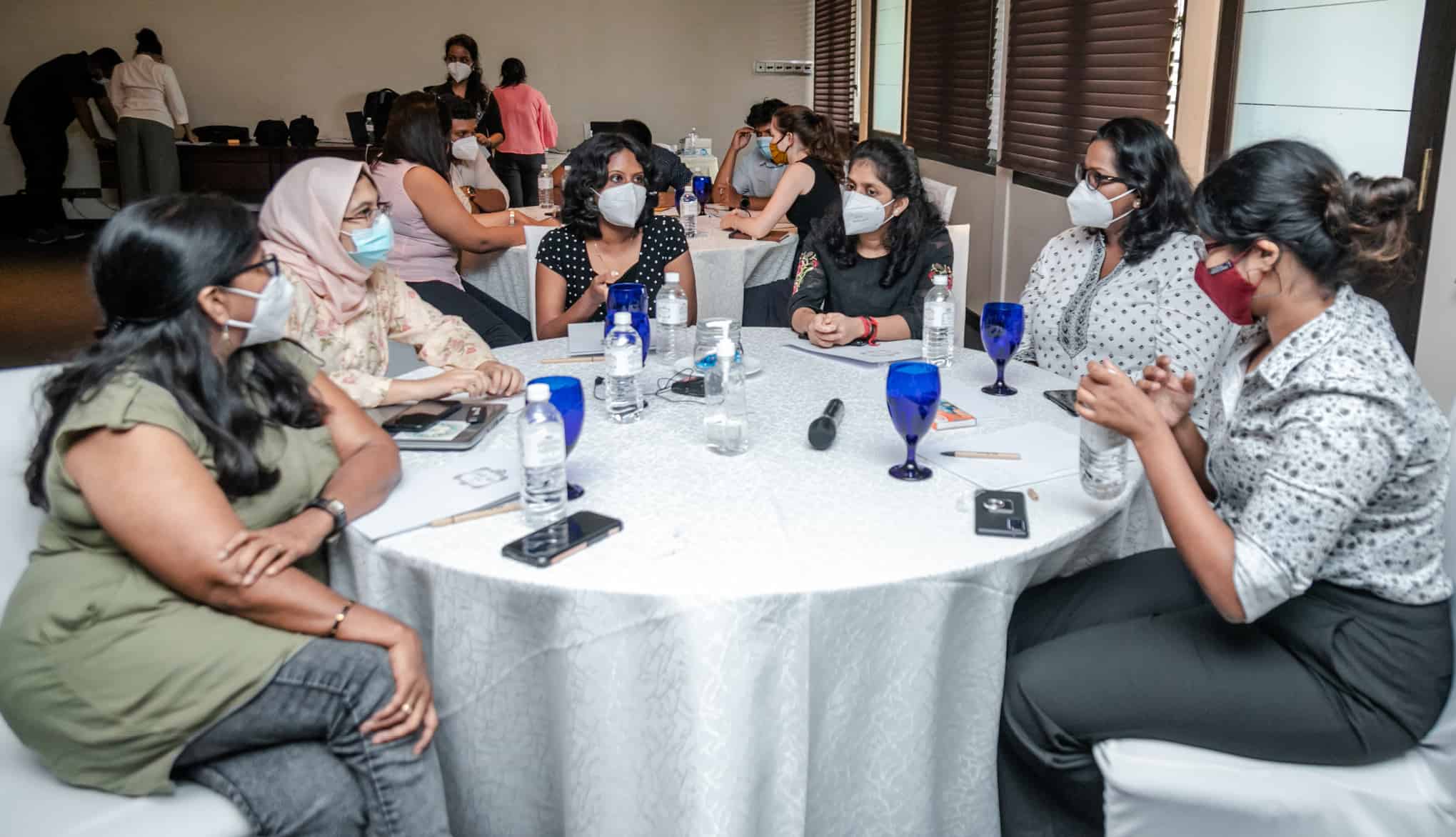 A group of women wearing face masks sit around a table talking.