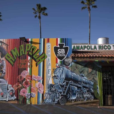 A mural is painted on a wall next to a restaurant called Amapola Rico Taco. The mural includes an old style car with two skeletons standing next to it and a steam engine train.