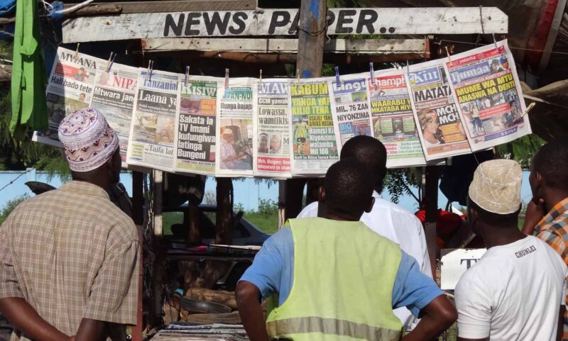 Three men read newspapers at a newstand.